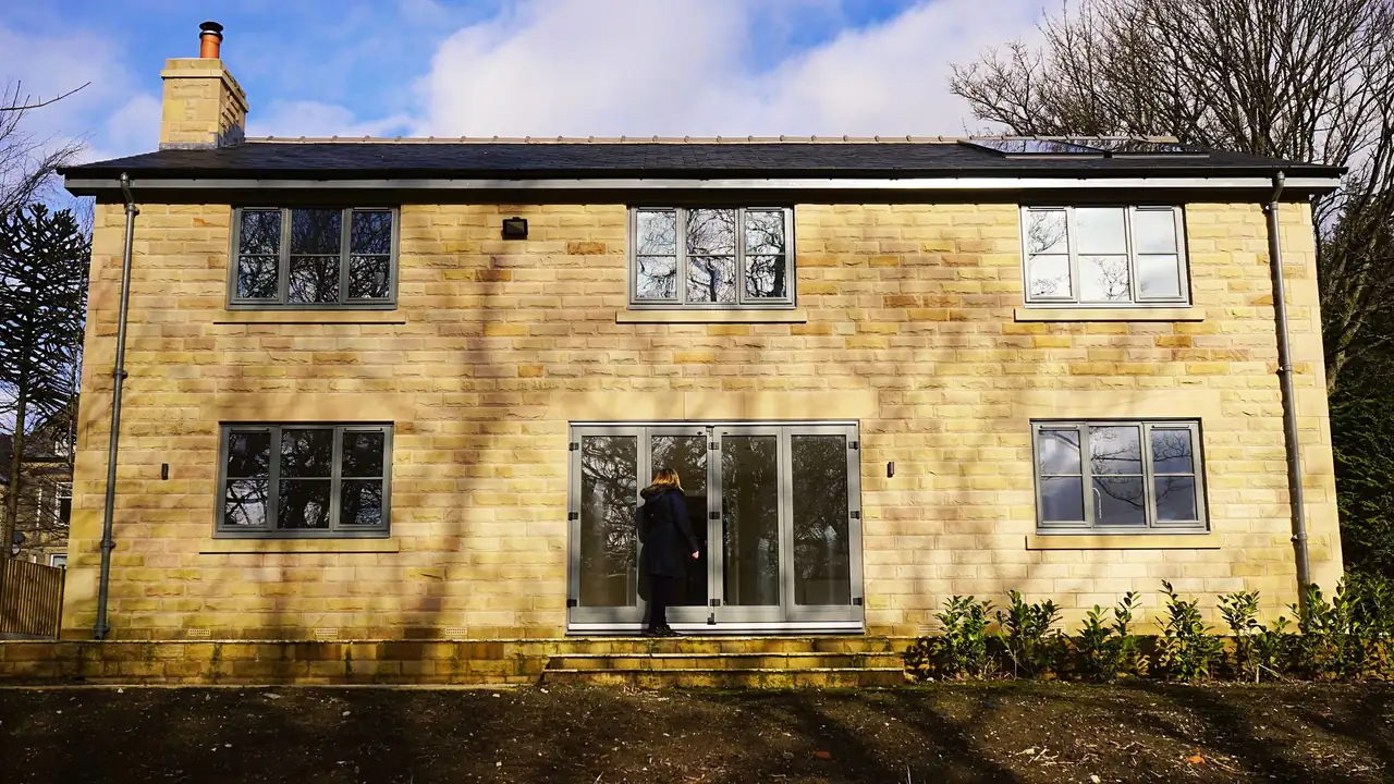 The back of a new two-storey house in honey-coloured cut stone, with grey-framed windows and bi-fold doors on to the garden