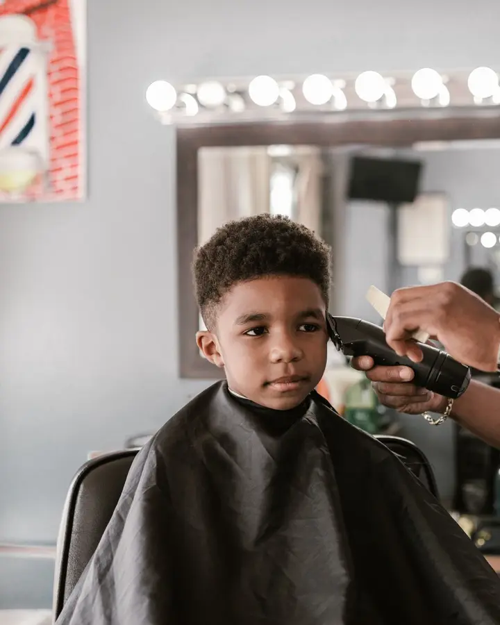 A young boy in a cape sitting still while a barber clippers the side of his head