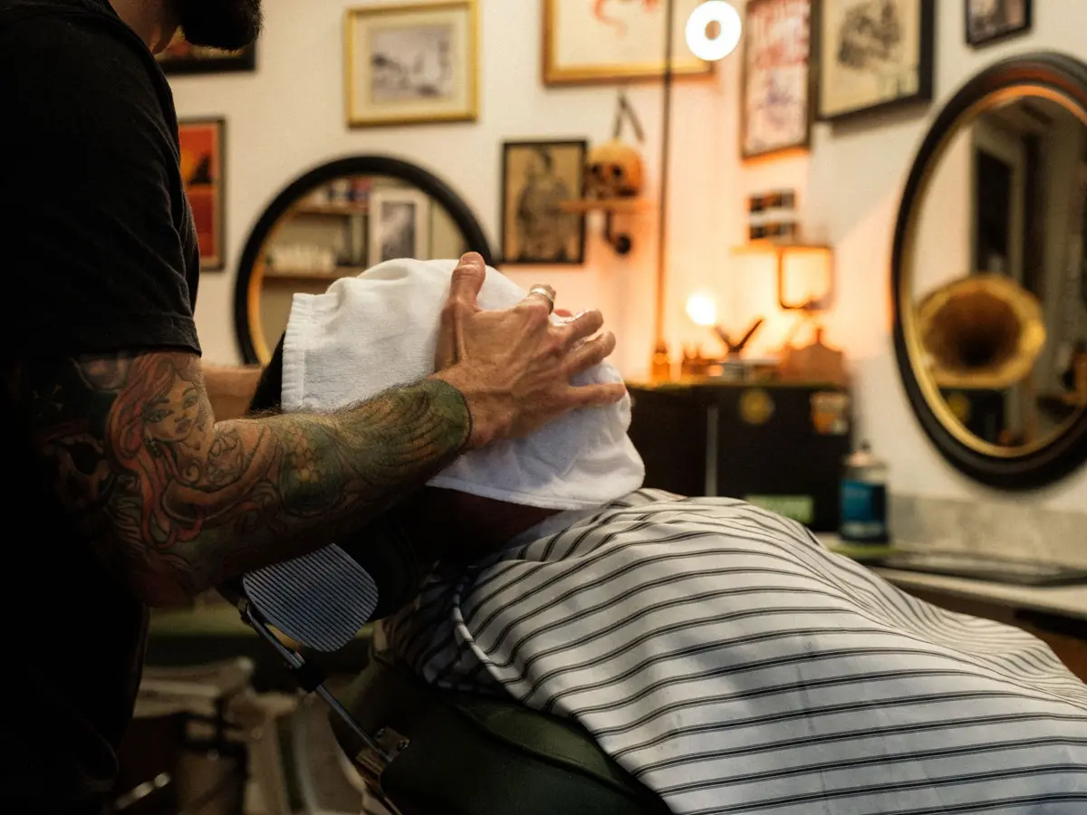 A tattooed barber pressing a hot towel over a reclined client's face in a vintage barbershop